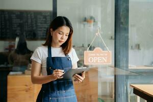 Young female hanging a welcome sign in front of a coffee shop. Beautiful waitress or hostess holding a tablet preparing  in a restaurant. photo