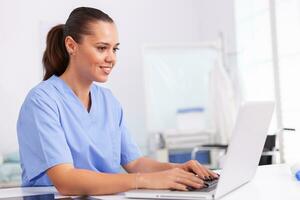 Medical nurse in uniform using laptop sitting at desk in hospital office. Health care physician using computer in modern clinic looking at monitor, medicine, profession, scrubs. photo