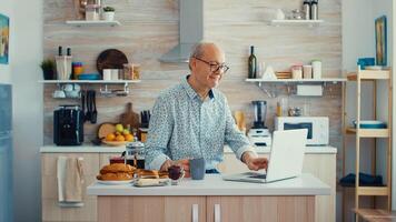 Daily life of senior man in kitchen during breakfast using laptop holding a cup of coffee. Elderly retired person working from home, telecommuting using remote internet job online communication on modern technology notebook photo