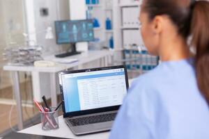 Medical nurse checking patient documentation on laptop in hospital office. Health care physician using computer in modern clinic looking at monitor, medicine, profession, scrubs. photo