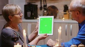 Old retired senior couple using green mockup screen digital isolated tablet computer. Aged people looking at green screen template chroma key display sitting at the table in kitchen during dinner. photo