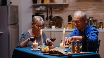 Portrait of senior couple with red wine glasses sitting at the table in the cozy kitchen. Happy cheerful elderly couple dining together at home, enjoying the meal, celebrating their anniversary photo