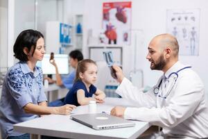 médico radiólogo explicando radiografía en hospital oficina durante médico cita. pediatra especialista hablando con madre acerca de enfermedad Proporcionar profesional cuidado de la salud tratamiento foto