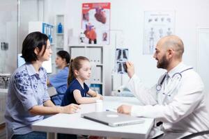 Pediatrician explaining diagnosis to mother and child holding radiography in hospital office. Healthcare physician specialist in medicine providing health care services treatment examination. photo