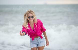 A beautiful woman in sunglasses and a pink shirt splashes sea water. photo