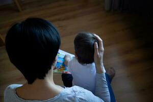 joven mamá con pequeño hijo leyendo libro mientras sentado en un silla en el dormitorio foto