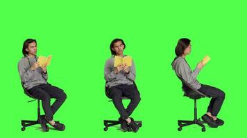 Asian guy reading literature book over greenscreen backdrop, enjoying lecture while he sits on chair in studio. Person reads fiction novel story, being interested in classic leisure activities. video