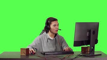 Asian guy sitting at office desktop, having fun with friends while enjoying internet multiplayer games with headphones. Over full body greenscreen, man competes in rpg contest. video