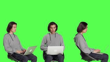 joven chico hojeada Internet en tableta en pantalla verde estudio, utilizando móvil artilugio a navegar mediante sitio web en línea. adulto sentado en silla y trabajando en dispositivo, relajado casual hombre. video