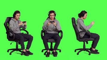 Asian guy sitting in chair over greenscreen, performing video games on mobile device. Young man utilizing phone to engage in smartphone games, web roleplaying contest with players.