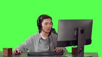 Male gamer at desk plays on computer, having fun with online multiplayer tournament for rpg video games with rivals. Asian guy gaming at pc over isolated greenscreen background.