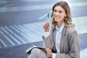 Business woman in suit sits in city centre, writes down, takes notes, holds pen and notebook, brainstorms, creates ideas photo