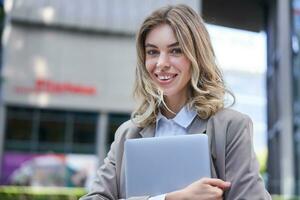 Portrait of young corporate woman, holding her laptop and smiling, going to work, wearing professional outfit, beige suite photo