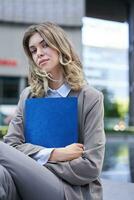 Corporate woman holding documents in hands, sitting on a street, wearing office suit, looking confident at camera, carry a folder with work photo