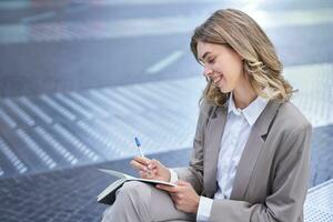 Business woman in suit sits in city centre, writes down, takes notes, holds pen and notebook, brainstorms, creates ideas photo