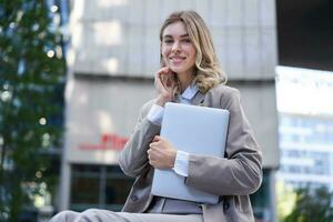 Portrait of young corporate woman, holding her laptop and smiling, going to work, wearing professional outfit, beige suite photo