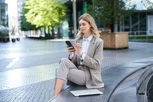 Business woman sits in city centre and works on mobile phone, she has her laptop and wears working corporate outfit photo