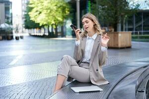 Business woman sits in city centre and works on mobile phone, she has her laptop and wears working corporate outfit photo