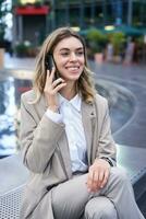 Vertical shot of smiling woman talking on mobile phone while sitting outside. Office lady waiting for someone after work photo
