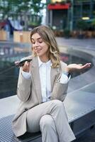 Vertical shot of smiling office lady in suit leaving voice message, talking into speaker on mobile phone, translating her speech on smartphone app photo