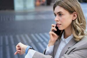 Busy lady manager talking on mobile phone on street, going on a meeting, checking time on watch photo