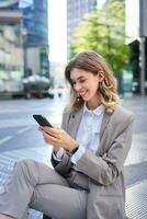 Vertical shot of smiling businesswoman looking at smartphone app, sending message on mobile phone, oder lunch while sitting outside office building photo