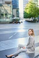 vertical Disparo de sonriente mujer de negocios en beige traje, sentado fuera de negocio oficina edificio, mirando con confianza foto