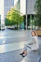 vertical Disparo de joven mujer de negocios en beige traje, giro alrededor, esperando para alguien fuera de oficina edificio foto