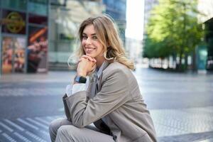 Portrait of woman waiting for an interview, sitting in city center in beige suit, has digital watch, smiling at camera photo