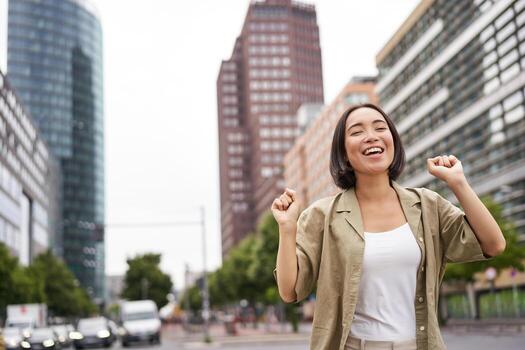 retrato de contento asiático mujer, bailando y sensación alegría, triunfando, levantamiento mano arriba en victoria gesto, celebrando en calles foto