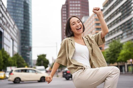 retrato de contento asiático mujer, bailando y sensación alegría, triunfando, levantamiento mano arriba en victoria gesto, celebrando en calles foto