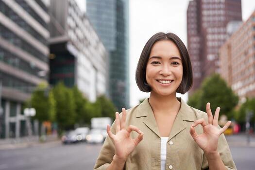 bueno, No problema. sonriente joven asiático mujer muestra Okay gesto, aprobar algo, me gusta y aceptar, aceptando, en pie en calle de ciudad foto