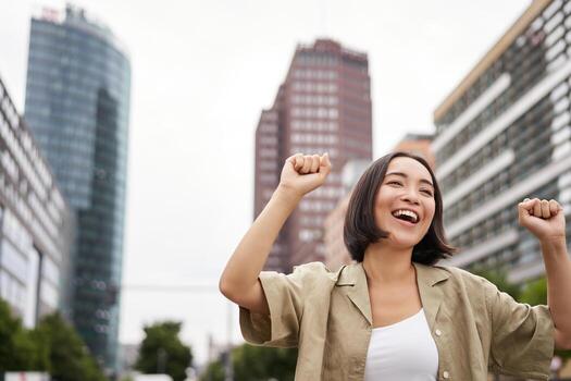 contento gente. joven sonriente asiático mujer bailando en ciudad, levantamiento manos arriba en ciudad, triunfando, celebrando victoria foto