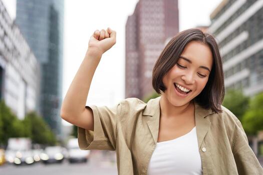 contento gente. joven sonriente asiático mujer bailando en ciudad, levantamiento manos arriba en ciudad, triunfando, celebrando victoria foto
