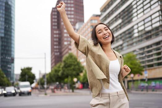 retrato de contento asiático mujer, bailando y sensación alegría, triunfando, levantamiento mano arriba en victoria gesto, celebrando en calles foto