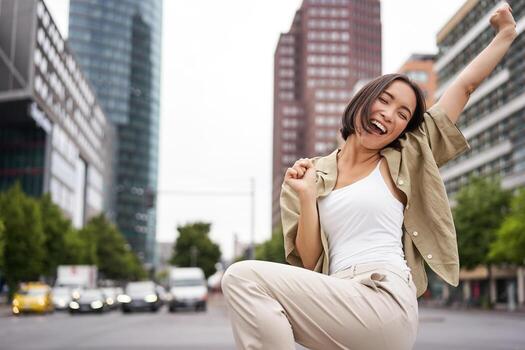 retrato de contento asiático mujer, bailando y sensación alegría, triunfando, levantamiento mano arriba en victoria gesto, celebrando en calles foto