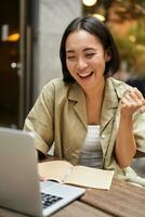 Vertical shot of happy girl talking on video call, looks at laptop, having online meeting, sitting in outdoor cafe photo