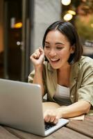 Vertical shot of happy girl talking on video call, looks at laptop, having online meeting, sitting in outdoor cafe photo