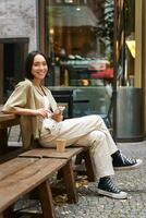 Portrait of stylish young korean woman sits in cafe, holds smartphone, smiles, enjoys coffee outdoors photo