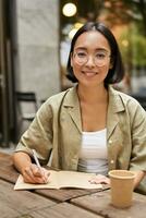 Vertical shot of young asian student, girl in glasses holding pen, making notes, writing in notebook and drinking coffee in cafe photo