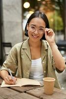 Girl studying from cafe, looking at camera while writing something in notebook, drinking coffee and smiling photo