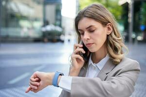 Serious businesswoman checking time on watch and calling person on mobile phone, waiting for someone in city photo