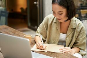 Portrait of young asian woman working on laptop, making notes, writing down while attending online lesson, work meeting photo