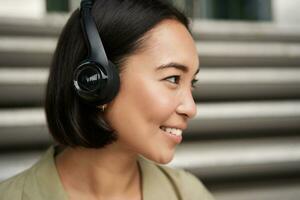Close up portrait of smiling asian girl in headphones, listens to music outdoors, looking happy. photo