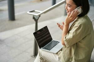 Smiling asian woman makes a phone call. Girl student using laptop and mobile phone, talking to someone on telephone photo