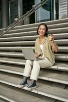 Vertical shot of smiling girl student, asian woman sits on stairs of university campus and drinks coffee, does her homework on laptop photo