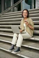 vertical Disparo de sonriente niña alumno, asiático mujer se sienta en escalera de Universidad instalaciones y bebidas café, lo hace su deberes en ordenador portátil foto