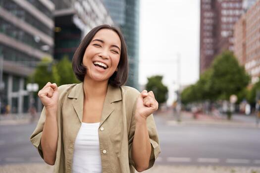 contento asiático niña triunfando en calles de ciudad, bailando desde felicidad, celebrando victoria, posando al aire libre foto