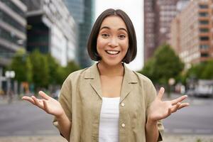 Modern asian girl standing on street with surprised face, looking impressed and amazed, hear awesome news photo