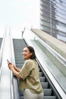 Young asian girl going up on an escalator, holding smartphone, smiling while walking in city photo
